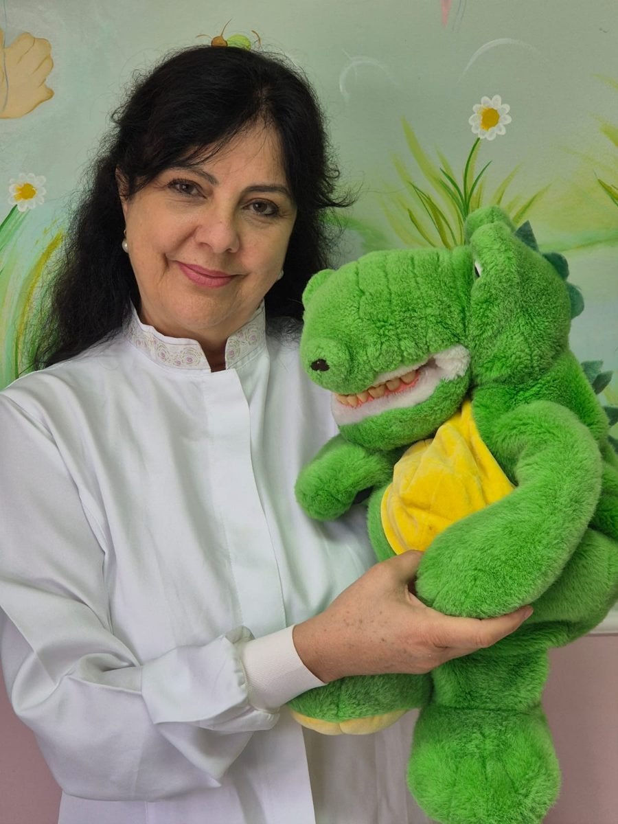 Dra. Marília Fernandes, de jaleco branco, sorrindo e segurando um jacaré de pelúcia usado nas consultas.