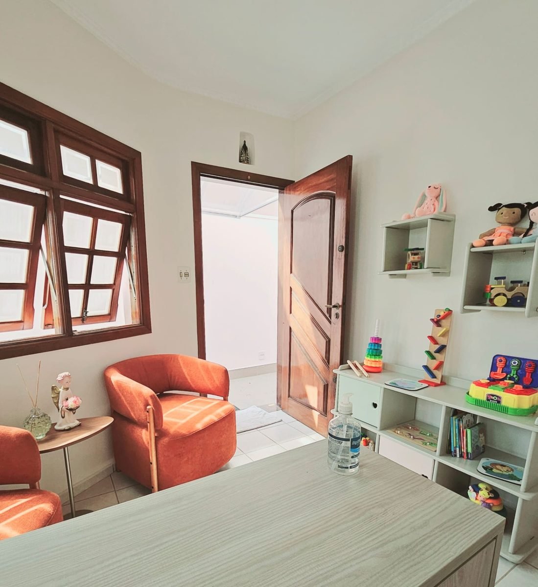 Sala de espera com poltronas laranja, janela de madeira e estante de brinquedos de madeira.