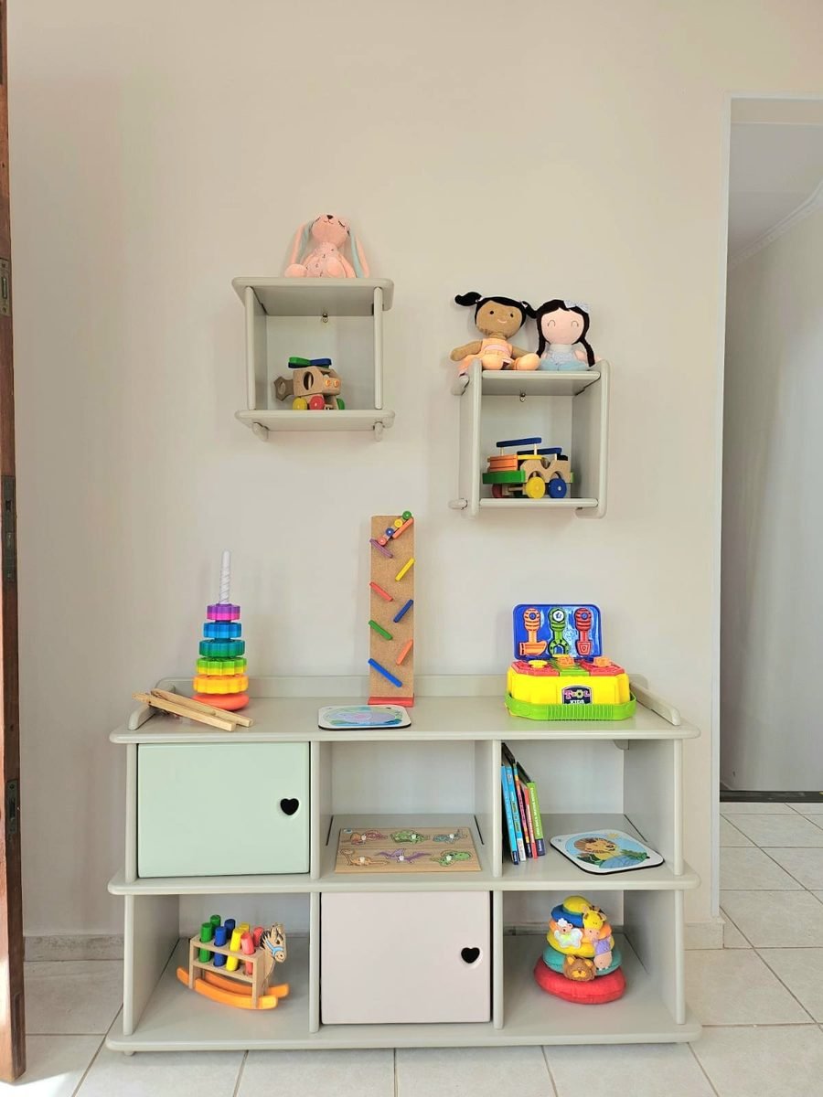 Estante clara com brinquedos de madeira, livros infantis, bonecas e quebra-cabeças.
