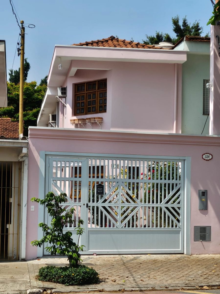 Fachada rosa do sobrado número 570, com portão branco e jardim na calçada.