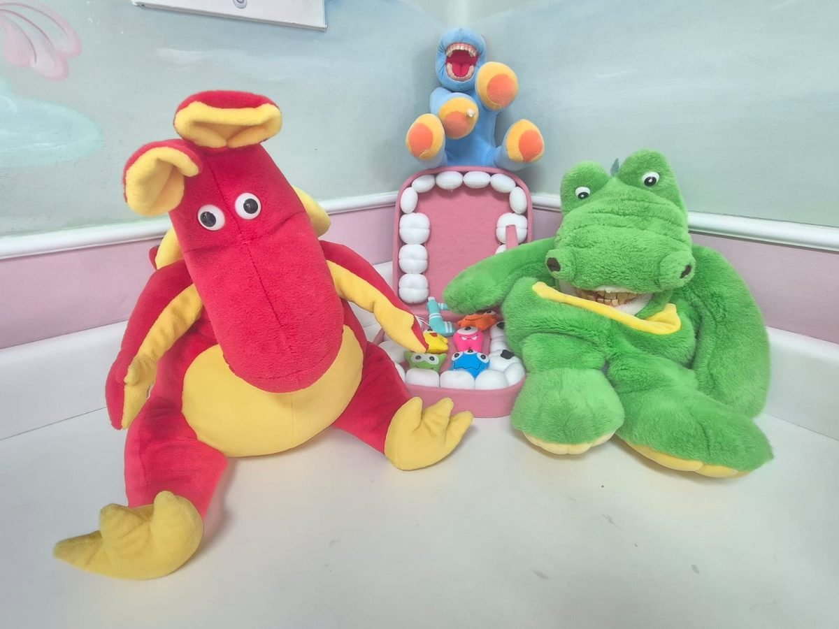 Pelúcias de dinossauro vermelho e jacaré verde com dentes, ao lado de uma boca de feltro rosa aberta.